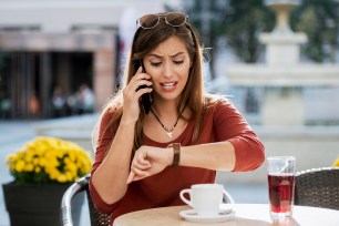 Young angry woman sitting in a cafe, drinking coffee and looking at her watch while making a phone call